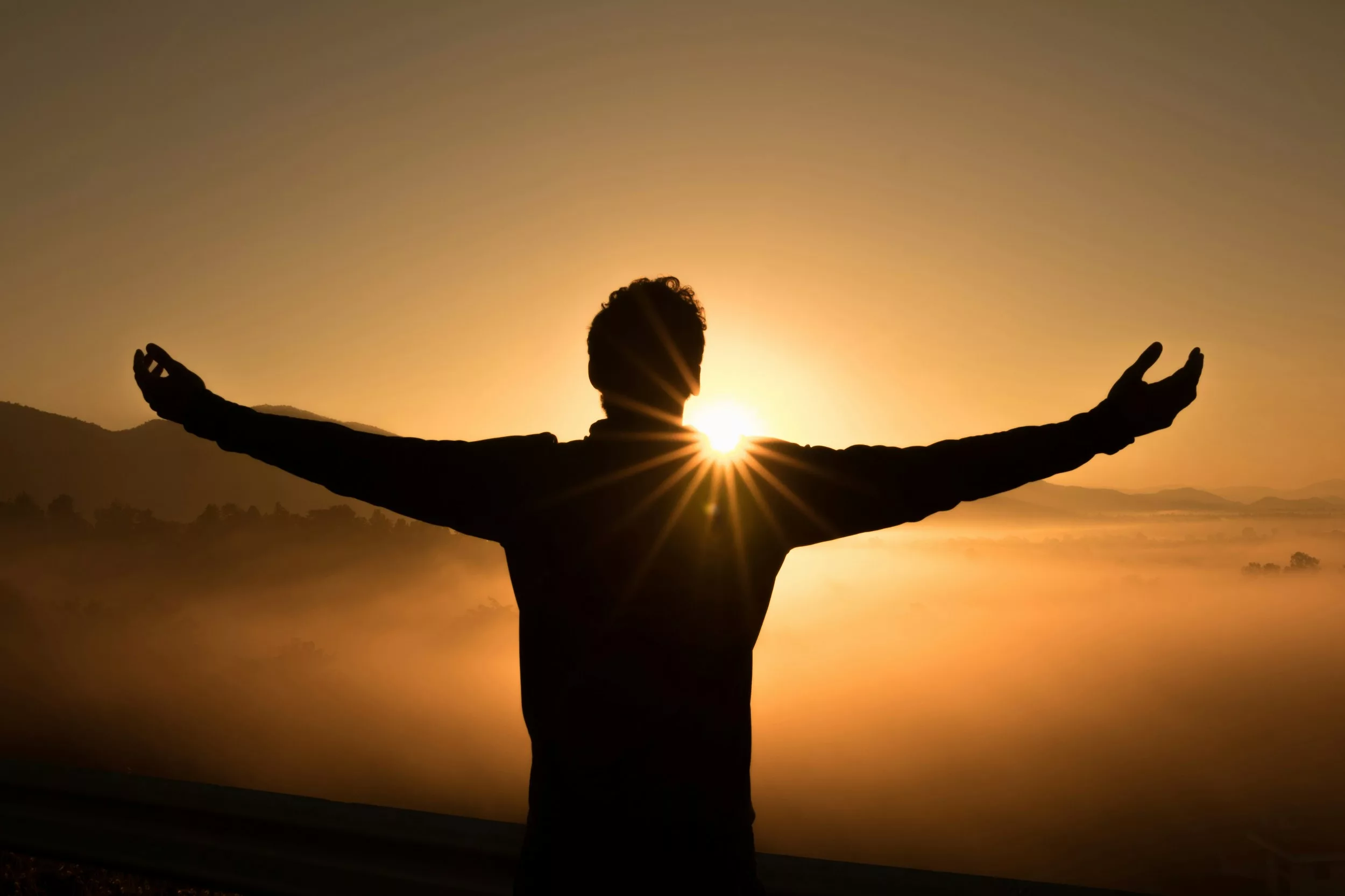 A person with arms open at sunrise over misty hills